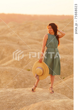 Young beautiful smiling woman with long hair in blue dress with straw hat in hand standing on sand in desert at sunrise 75668023