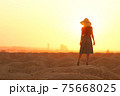 Elegant woman in long dress with straw hat standing on sand in desert, back view. Sunrise, backlight photo 75668025