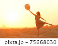 Elegant woman with long hair in dress standing on one leg in sandy desert, straw hat raised in hand, sunrise, backlight 75668030