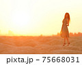 Beautiful woman with long hair in blue dress with straw hat in hand standing on sand in desert at sunrise, backlight 75668031