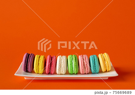 Multicolored macaroons in a row on a white rectangular plate against bright red background. Multicolored macaroons in a row on a white rectangular plate against bright red background. 75669689