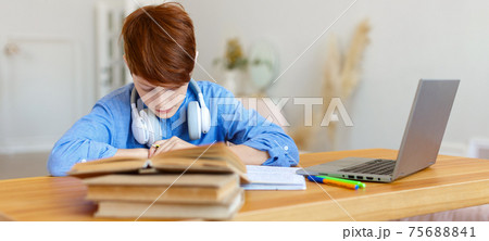 A pupil boy writes an assignment in a notebook during an online lesson in front of a laptop monitor. A pupil boy writes an assignment in a notebook during an online lesson in front of a laptop monitor. 75688841
