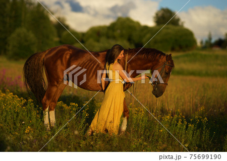 Beautiful woman with long hair in yellow dress standing near brown horse in among purple flowers in green field Beautiful woman with long hair in yellow dress standing near brown horse in among purple flowers in green field 75699190