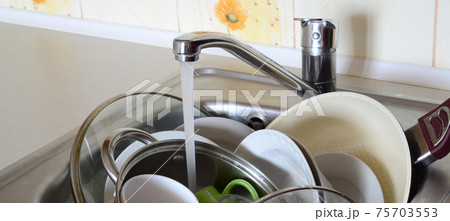 Dirty dishes and unwashed kitchen appliances lie in foam water under a tap from a kitchen faucet 75703553
