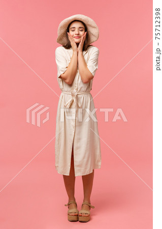 Full-length vertical portrait happy cheerful woman feeling uplifted and dreamy, close eyes smiling touching face, moisturizer walking on sunny perfect day, standing pink background 75712398