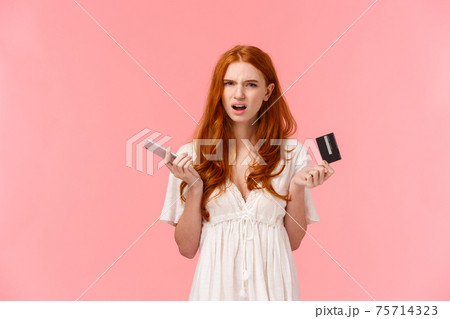 Frustrated and annoyed redhead girl feel displeased with strange online banking system, cant get clue what happened her order, holding smartphone and credit card squinting bothered 75714323