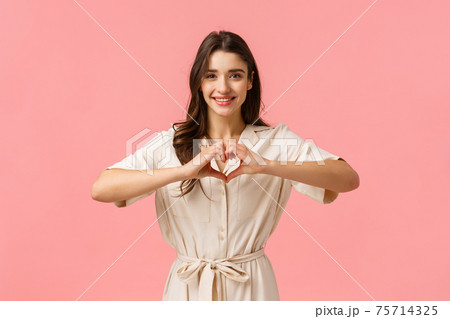 Waist-up portrait charming young brunette girl in dress, showing heart sign with love or care, absolutely adore boyfriend, having perfect relationship, feeling happy, smiling joyfully 75714325
