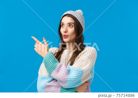 Girls attention caught something interesting happening nearby. Intrigued, curious good-looking brunette woman in winter sweater, hat, looking pointing left with folded lips, amazed, blue background Girls attention caught something interesting happening nearby. Intrigued, curious good-looking brunette woman in winter sweater, hat, looking pointing left with folded lips, amazed, blue background 75714886