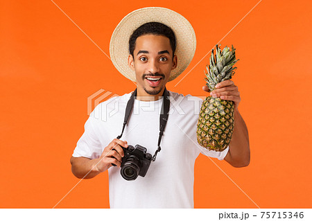 Charismatic young attractive african-american tourist, traveller taking pictures from vacation on camera, holding pineapple and smiling joyfully, having fun on summer luxury resort, orange background 75715346