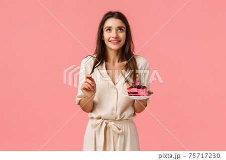 Dremy cute, feminine brunette female holding spoon and plate, dreaming, smiling while looking up and making wish blowing-out candle on birthday cake, having b-day party, celebrating, pink background 75717230