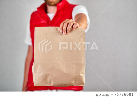Courier, delivery man in red vest uniform delivers online purchases in brown paper bags 75726081