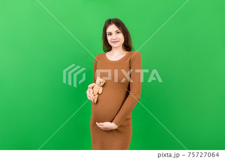 Happy pregnant woman holding a teddy bear against her belly at Colored background. Young mother is expecting a baby. Copy space Happy pregnant woman holding a teddy bear against her belly at Colored background. Young mother is expecting a baby. Copy space 75727064
