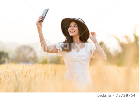 Happy young beautiful woman wearing black hat and white dress taking selfie while walking in the golden barley filed on a late afternoon Happy young beautiful woman wearing black hat and white dress taking selfie while walking in the golden barley filed on a late afternoon 75729754