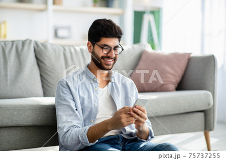 Arab guy texting and browsing internet on smartphone, resting at home, leaning on sofa Arab guy texting and browsing internet on smartphone, resting at home, leaning on sofa 75737255