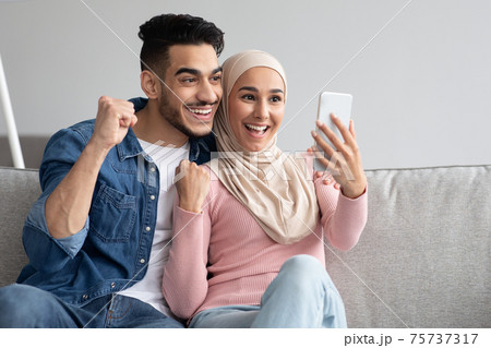 Cheerful arab couple with smartphone raising fists up 75737317