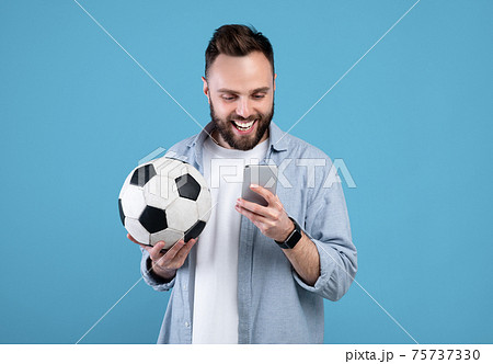 Happy young guy with soccer ball using smartphone, winning sports bet, rooting for his favorite team on blue background 75737330