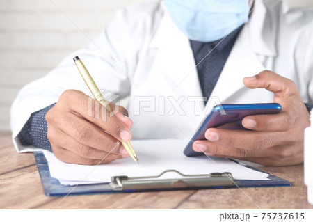doctor in face mask writing prescription and using smart phone  75737615
