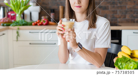 Happy brunette woman sitting with homemade glass smoothie and healthy fruits at home kitchen. Vegan meal and detox concept. Girl with white t-shirt drinking fresh cocktail 75738505