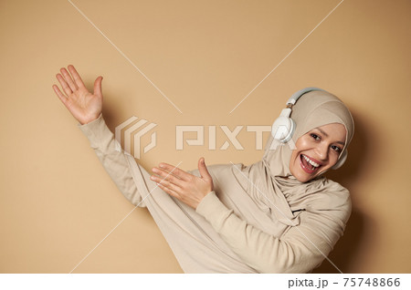 Gorgeous Muslim woman in hijab listening to the music in headphones, pointing hands on beige background with copy space 75748866