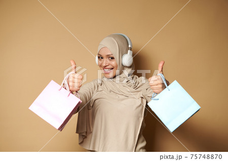 Attractive woman in hijab and headphones holding pink and blue gift paper bags in the hands and showing thumbs up, expressing happiness of upcoming religious holidays 75748870