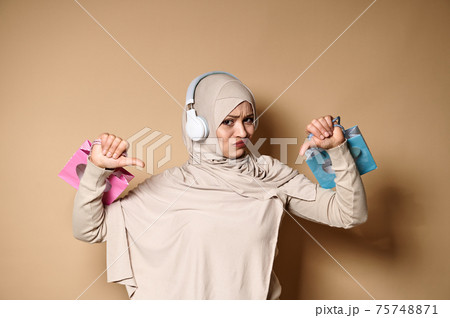 Upset woman in hijab and headphones holding color gift paper bags in the hands and showing thumbs down, expressing dissatisfaction 75748871