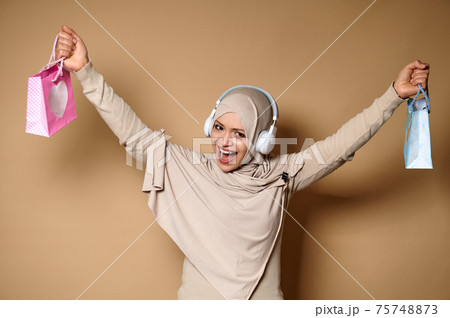 Smiling Arab Muslim woman in hijab and headphones holding color paper bags and expressing happiness looking at the camera. 75748873