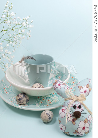 Easter festive composition for holiday breakfast with beautiful tableware, quail eggs and DIY textile Easter bunny on light blue backdrop. Copy space for text. Vertical orientation. 75766743