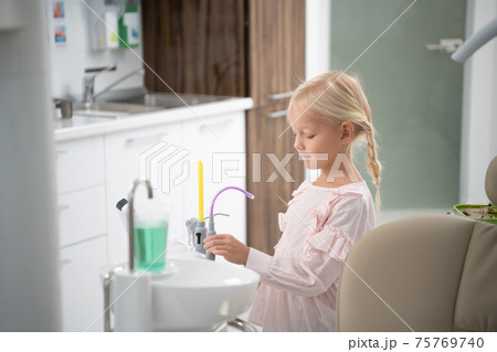 A little blond girl in the dental clinic. A cup with sterile water for rinsing the mouth in the foreground. A visit to a dentist concept 75769740