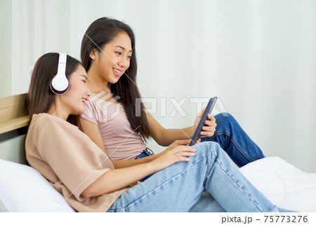 Young asian teen and her friend use tablet computer on the bed to relax in the bedroom, enjoy leisure weekend at home. Stress free concept 75773276