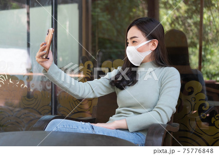 Young woman wearing medical mask making selfie or video call with smartphone at coffee shop. Young woman wearing medical mask making selfie or video call with smartphone at coffee shop. 75776848