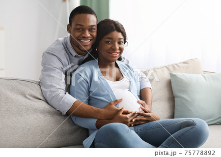 Smiling black man hugging his pregnant woman sitting on sofa 75778892
