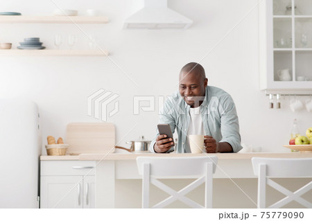 Happy african american man using smartphone and having morning coffee in the kitchen Happy african american man using smartphone and having morning coffee in the kitchen 75779090