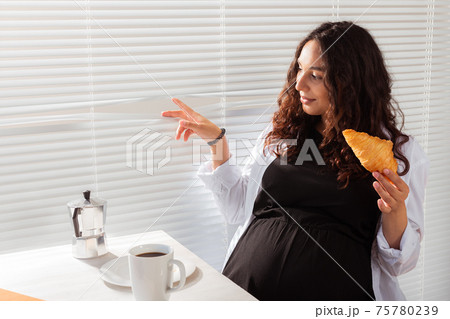 Happy pregnant young beautiful woman eating croissant and looking througt blinds during morning breakfast. Concept of pleasant morning and positive attitude during pregnancy Happy pregnant young beautiful woman eating croissant and looking througt blinds during morning breakfast. Concept of pleasant morning and positive attitude during pregnancy 75780239
