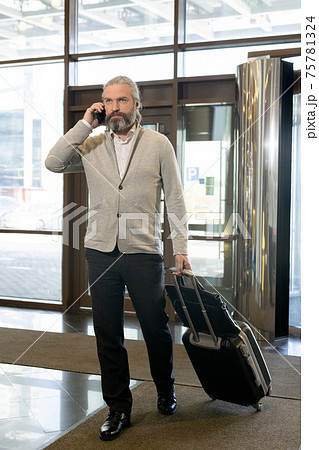 Mature businessman with smartphone by ear pulling suitcase while entering hall of hotel Mature businessman with smartphone by ear pulling suitcase while entering hall of hotel 75781324