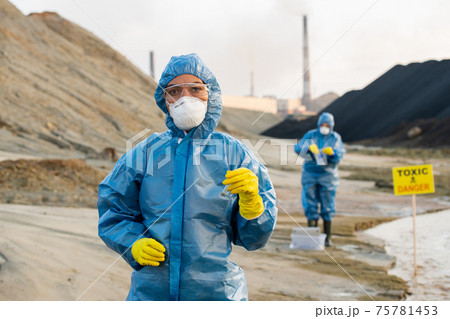 Young serious female researcher in protective workwear holding sample of toxic soil in flask 75781453