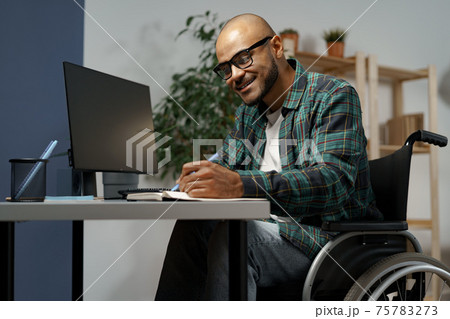 Disabled young african american man in wheelchair using computer while sitting at his working table Disabled young african american man in wheelchair using computer while sitting at his working table 75783273