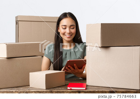 Small business owners, startup and e-commerce concept. Successful asian businesswoman work home, manage internet store from digital tablet, packing orders for shipping, sitting with piles of boxes 75784663
