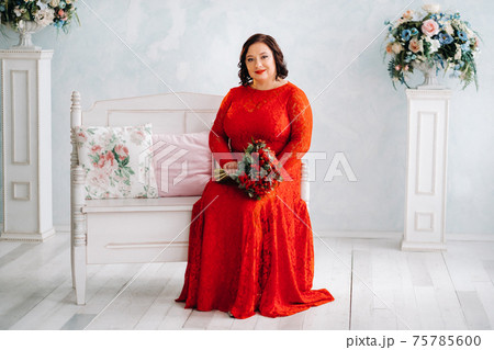 a woman in a red dress sits on a sofa and holds a bouquet of red roses and strawberries in the interior. a woman in a red dress sits on a sofa and holds a bouquet of red roses and strawberries in the interior. 75785600