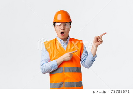 Building sector and industrial workers concept. Asonished and impressed asian male architect, engineer in protective helmet and safety glasses working at factory, pointing upper right corner amazed Building sector and industrial workers concept. Asonished and impressed asian male architect, engineer in protective helmet and safety glasses working at factory, pointing upper right corner amazed 75785671