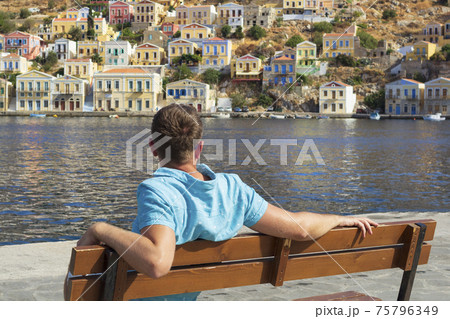 Young male tourist sitting on a bench and looking at the port of Symi, Greece with many different color houses. 75796349