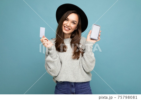 Beautiful smiling young brunet woman wearing black hat and grey sweater isolated over blue Beautiful smiling young brunet woman wearing black hat and grey sweater isolated over blue 75798081