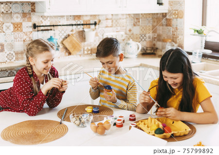 Two sisters and a brother are sitting at the table and painting Easter eggs in different colors for Easter, laughing and indulging 75799892