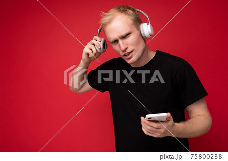 Upset asking handsome blonde young man wearing black t-shirt and white headphones standing isolated 75800238
