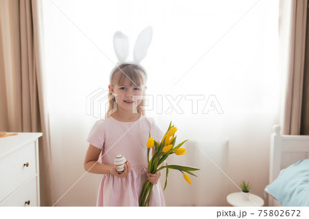 Easter concept. Girl in bunny ears and pink dress holding yellow tulips and egg Easter concept. Girl in bunny ears and pink dress holding yellow tulips and egg 75802672