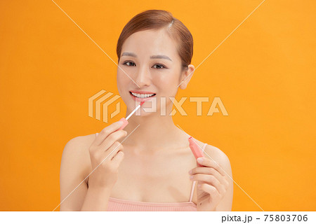 Beauty portrait of a pretty young topless woman wearing makeup isolated over yellow background, applying lip gloss 75803706