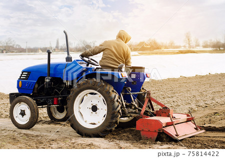Farmer on a tractor with milling machine loosens, grinds and mixes soil. Loosening the surface, cultivating the land for further planting. Farming and agriculture. Cultivates the soil. Plows a field. 75814422