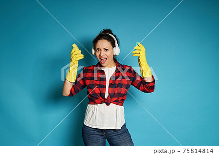 Young african woman in red plaid shirt with headphones on her head sings, posing on a blue background with cleaning sponges in her hands Young african woman in red plaid shirt with headphones on her head sings, posing on a blue background with cleaning sponges in her hands 75814814