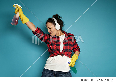 Hispanic woman, housewife, with headphones holding cleaning sprays in her hands and dancing with her eyes closed, enjoying her homework. Hispanic woman, housewife, with headphones holding cleaning sprays in her hands and dancing with her eyes closed, enjoying her homework. 75814817