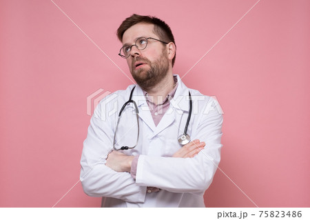 Tired male doctor in a white coat, with a stethoscope around neck, crossed arms and looked up 75823486