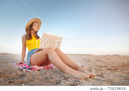 Young Caucasian Woman in shorts and a hat on the beach sitting on the sand browses social networks on the computer in the summer against the background of the sea. Remote work with a laptop 75829658